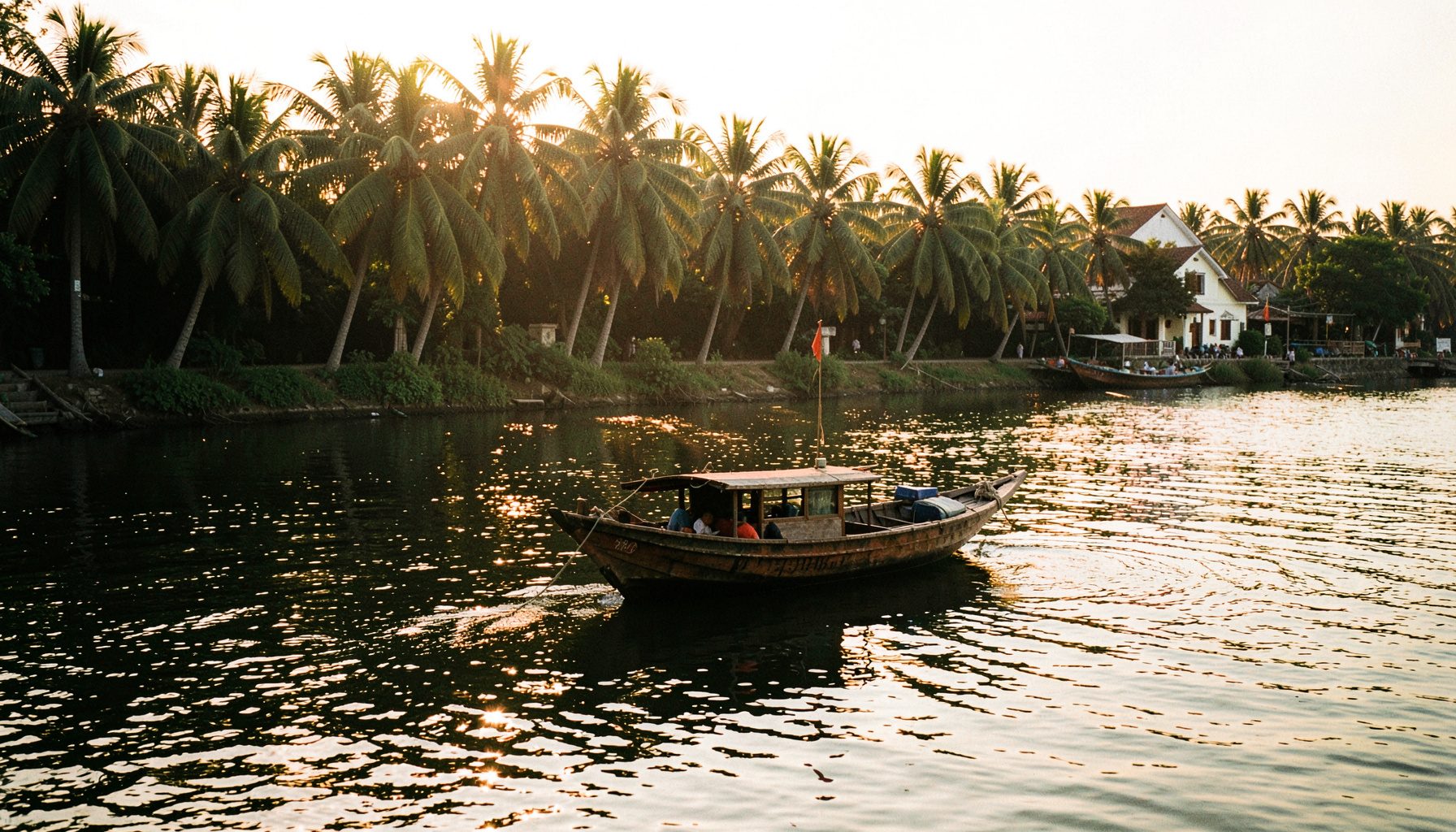 A sunlit wooden boat drifting on calm river water in Hoi An, with lush green palm trees along the bank and a warm golden sky at dusk