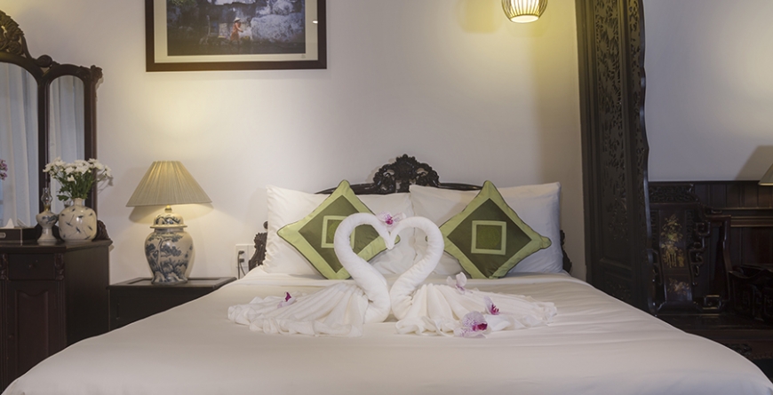 Bed with white linens, green patterned pillows, and towels folded into a swan shape, soft warm lighting, romantic mood