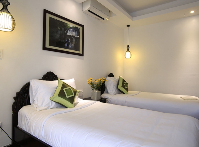 Twin bedroom with white linens, green accent pillows, dark carved headboards, framed artwork on wall, soft warm lighting