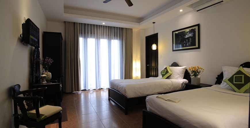 Hotel bedroom with two twin beds with white linens and green patterned pillows, dark wood furniture, curtained balcony doors