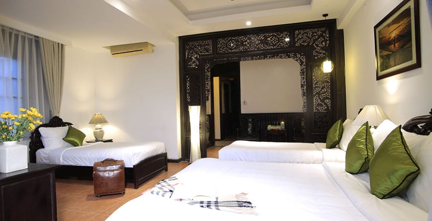 Hotel bedroom with multiple beds with white linens and green pillows, ornate dark carved wood partition, warm lighting