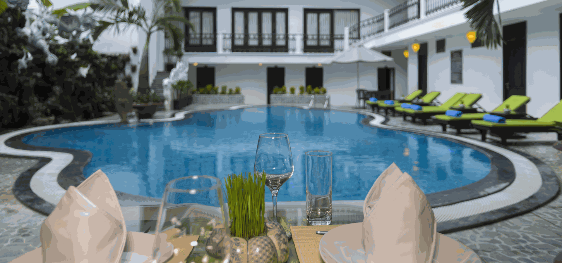 Outdoor dining table with glasses and napkins in the foreground, a curved swimming pool and lounge chairs behind, under a bright blue sky with white and green tones