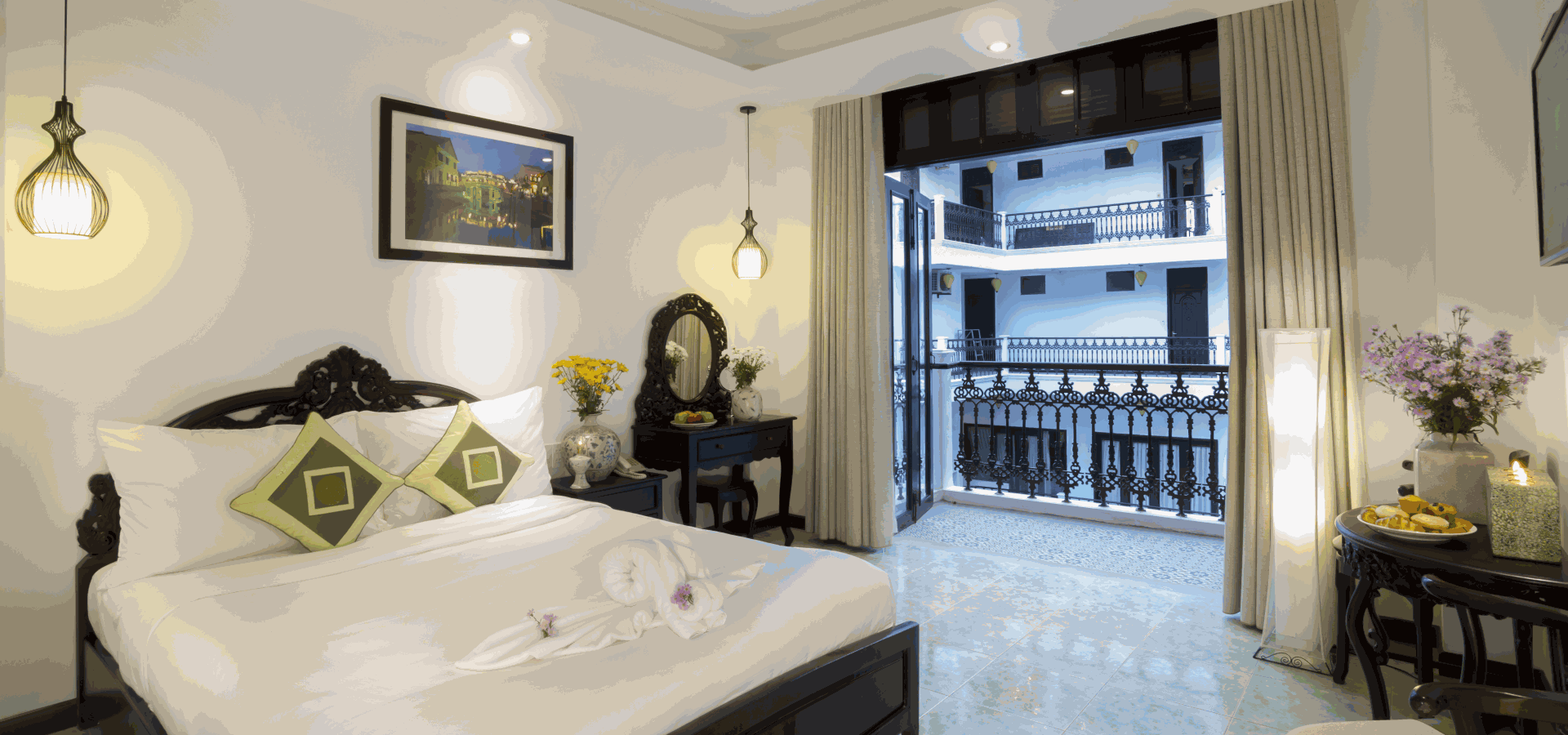 Hotel bedroom with dark wood bed frame, white linens, green accent pillows, flowers, and a view of balconies through open doors