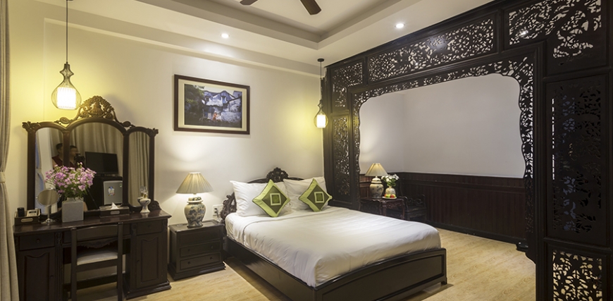 Hotel bedroom with a bed with white linens and green pillows, ornate dark carved wood canopy frame, vanity with mirror