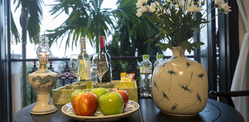 A warmly lit restaurant table set with a bowl of fresh apples, decorative vases with flowers, wine bottles and glasses, and water bottles, with greenery in the background